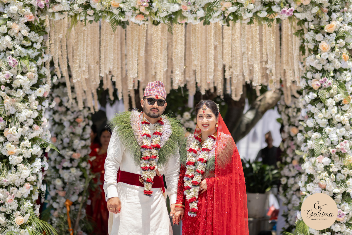 Nepali bride and groom standing beneath a white floral wedding arch 