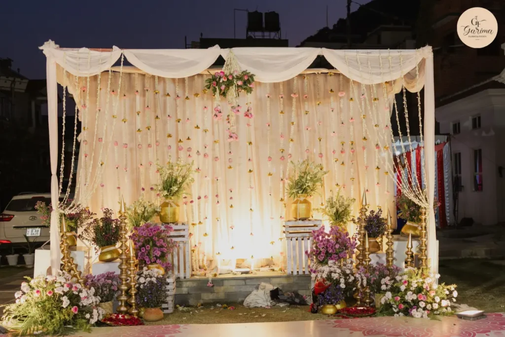 simple white draped haldi backdrop with hanging flowers and floral arrangements